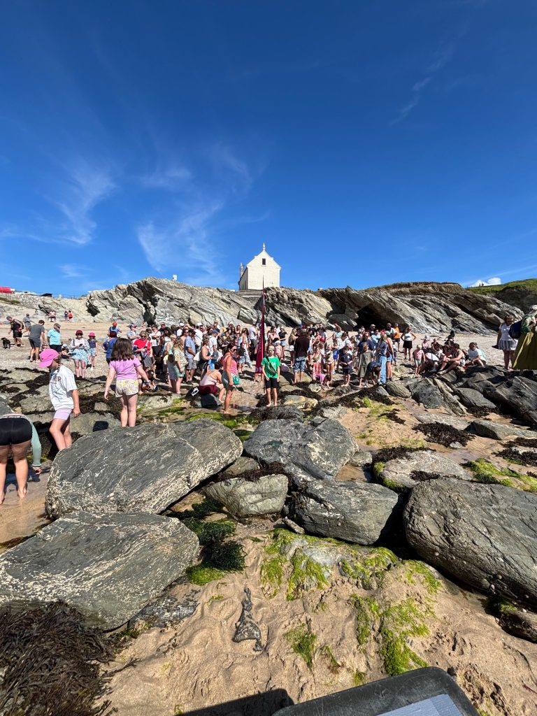 Image showing people gathered for the talk by the National Lobster Hatchery Team. From the perspective of the shoreline looking into the beahc, with the lifeboat house and blue sky in the background.